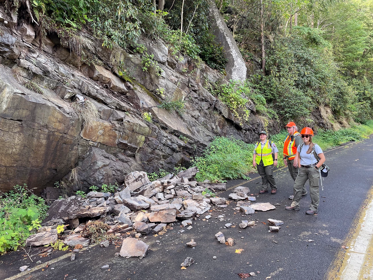 Park staff at a rockfall site Three people in hard hats and safety vests stand on a road beside a rock fall, where part of a rocky cliff backing the road has crumbled onto it.
