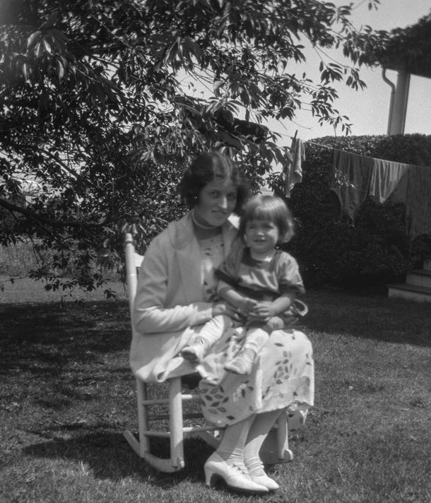 A black and white photo of a woman in a white dress, jacket, and heeled shoes sitting in a rocking chair with a small child on her lap, on a grassy lawn in front of a partly visible cottage, tree, and clothes drying on a line.