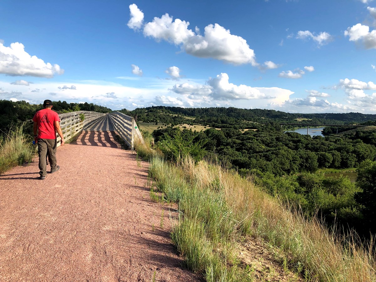 Man in red shirt walks on trail heading onto a foot bridge across a river. Blue sky with clouds overhead, in the distance another bridge is in view crossing the river upstream