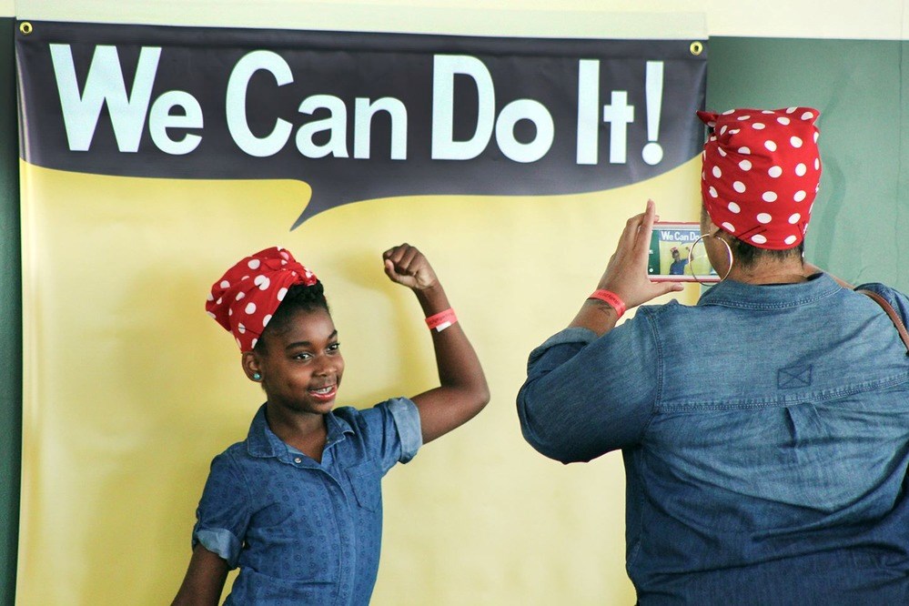 National Park Service. Rosie the Riveter National Historic Park. Color photo of a young African American girl in blue coveralls and red bandana with white polk dots having her picture taken by an adult in blue coveralls with matching bandana in front of a speechbubble reading "We Can Do It!"