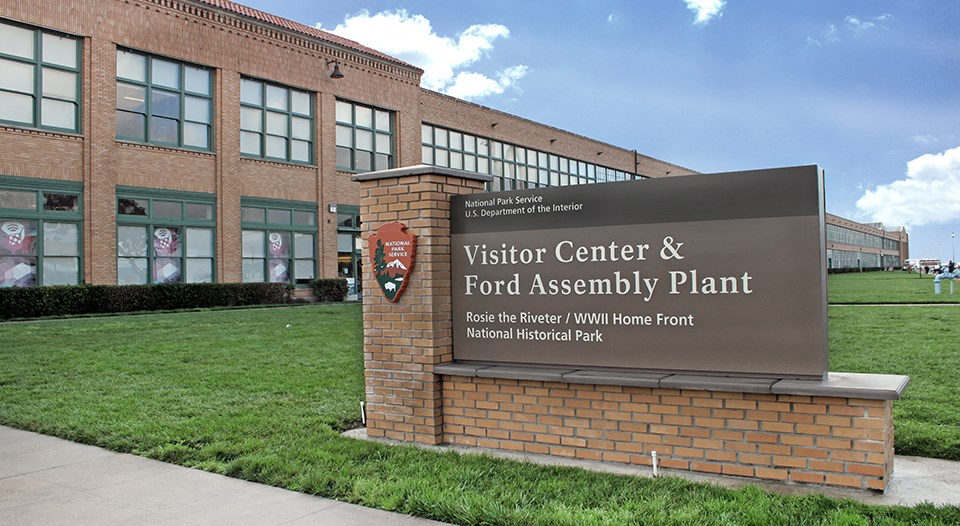 a park sign in front of a two-story industrial building says Visitor Center, Ford Assembly Plant