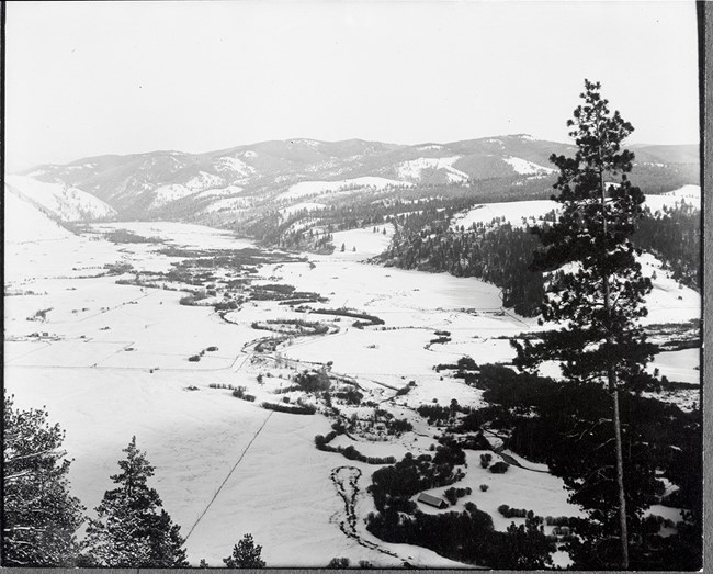 Black and white aerial photograph of a snow-covered landscape with mountains in the background and trees in the foreground.
