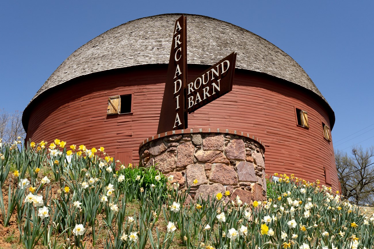 A round building with a rounded roof, surrounded by flowers. A sign reads "Arcadia Round Barn". A round building with a rounded roof, surrounded by flowers. A sign reads "Arcadia Round Barn".