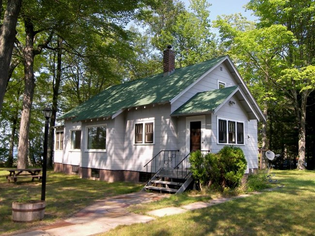 White house with green pointed roof and brick chimney, surrounded by trees.