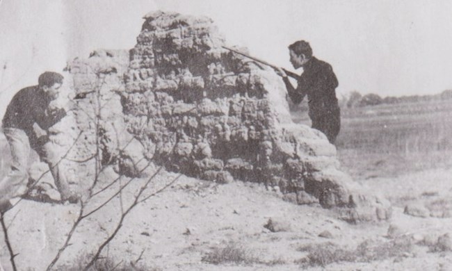 Two men with rifles play around a ruined adobe wall