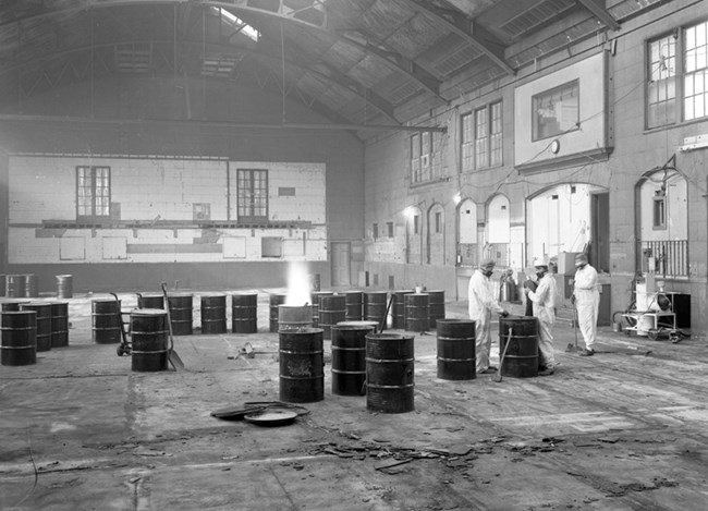 Runnymede, Unit 4, Demolition and Clean-up 3 people wearing respirators and protective clothing work in large gymnasium that is empty except for 20 50-gallon metal drums. A fire burns in one of the drums.