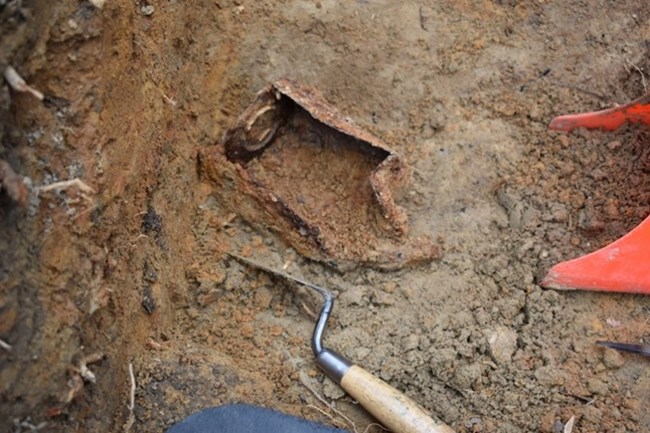 A rusty tin can sitting in the middle of an open dirt unit with a metal trowel and red plastic shovel near it.