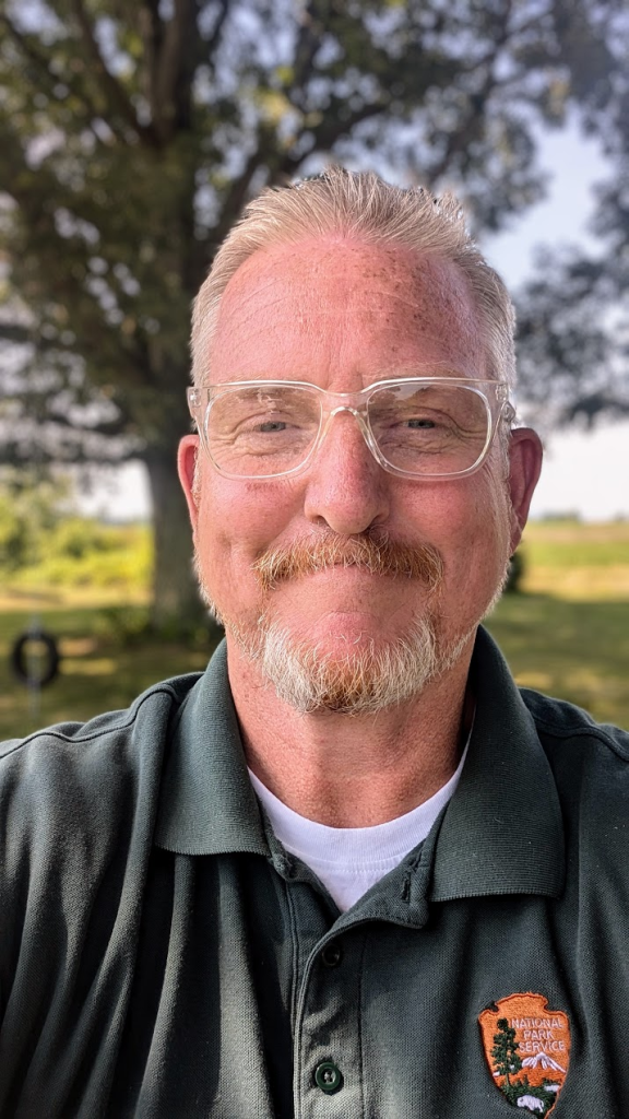 A smiling man with short blonde hair and a trimmed beard, wearing clear glasses and a dark green polo shirt with the National Park Service logo. He is standing outdoors with a tree and blurred greenery in the background, under daylight conditions. A smiling man with short blonde hair and a trimmed beard, wearing clear glasses and a dark green polo shirt with the National Park Service logo. He is standing outdoors with a tree and blurred greenery in the background, under daylight conditions.