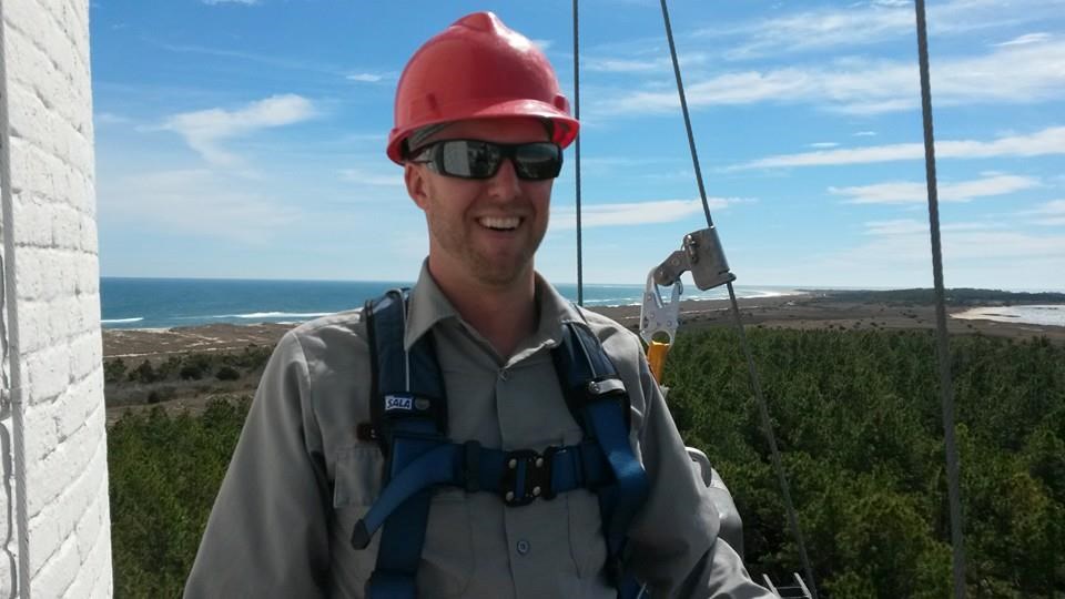 Cape Lookout Lighthouse Exterior Painting Sabol wears a hardhat, glasses and harness on the Cape Lookout Lighthouse with trees and the sea in the background below him.