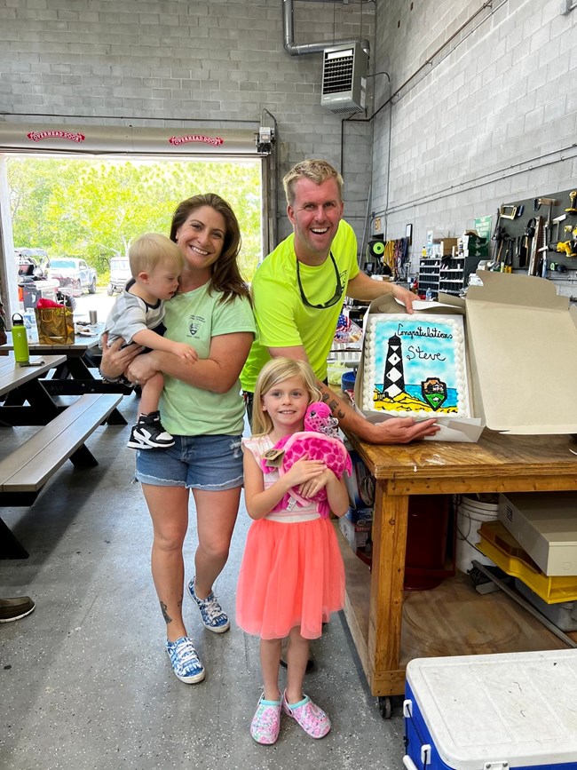 Steve Sabol Cape Lookout Farewell Steve stands inside a building next to family holding up a farewell cake.