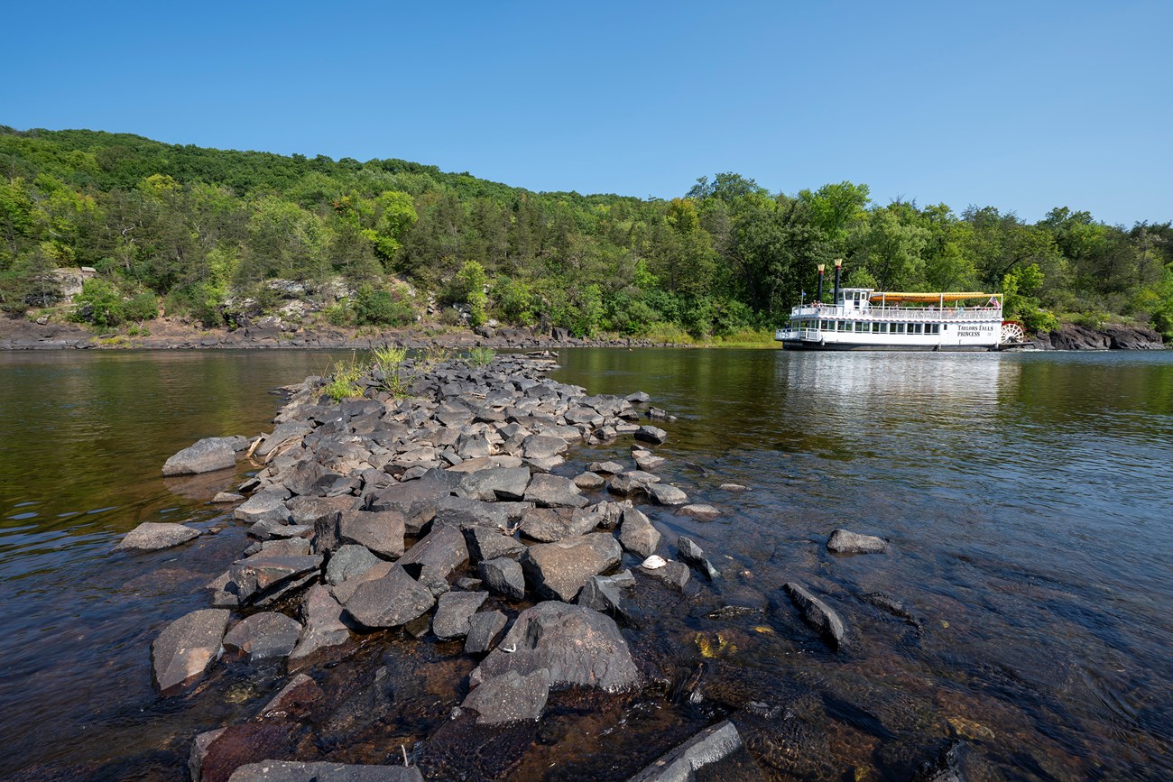 a paddlewheel riverboat travels past a historic closing dam that appears as a pile of rocks crossing the river