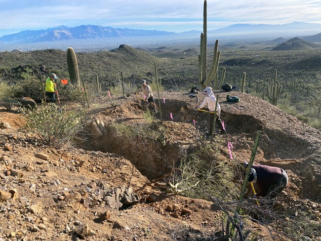 A crew of four NPS workers using tools to fill a large opening of an abandoned mine site. There are mountains and cacti in the landscape.