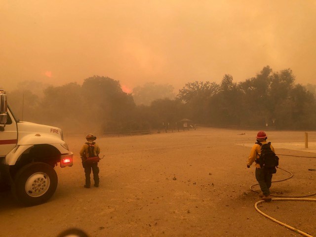 Two firefighters face a group of trees, with so much smoke that the whole picture is tinted orange.