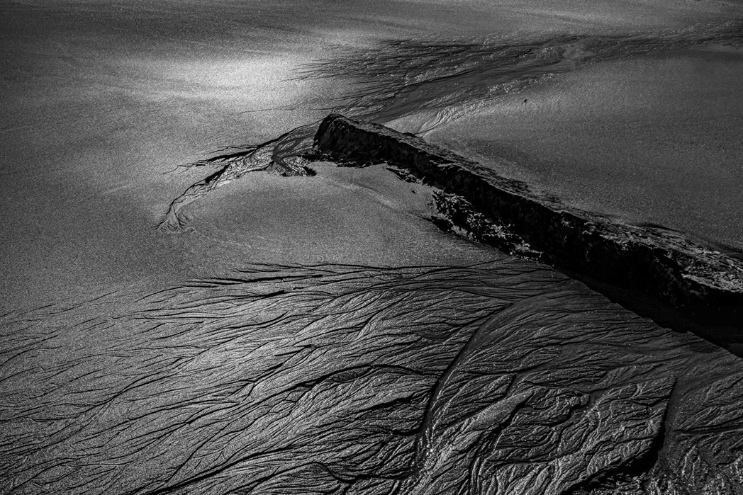 High contrast black and white photograph of wet sand intersected with a rectangular stone and fine, interlaced ripples tracing across the surface