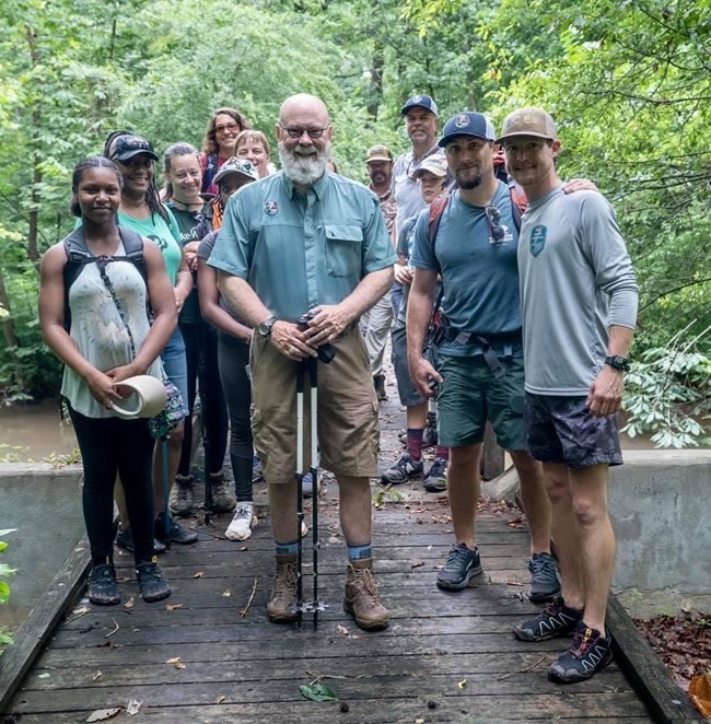 A group of people stand smiling in hiking gear crossing small wooden bridge in woods
