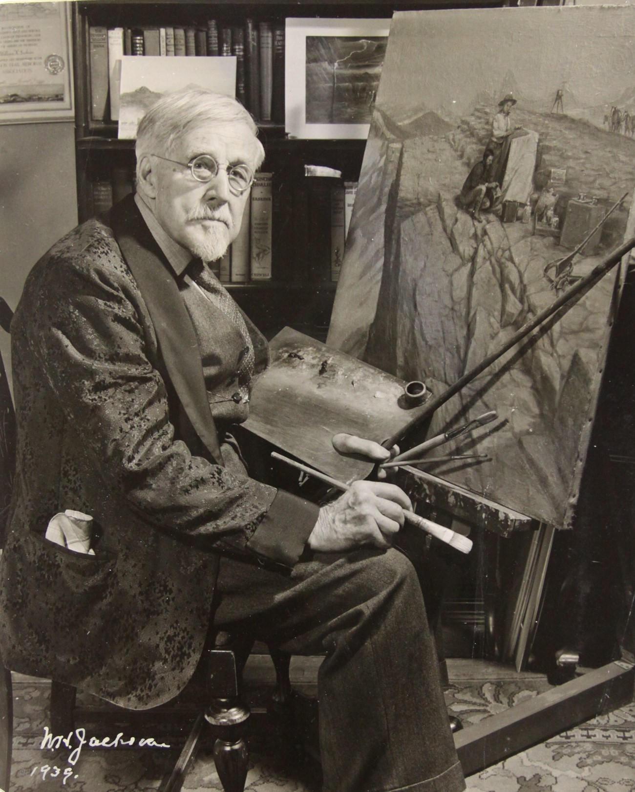An old man sits at an easel holdig a palette and paintbrush.
