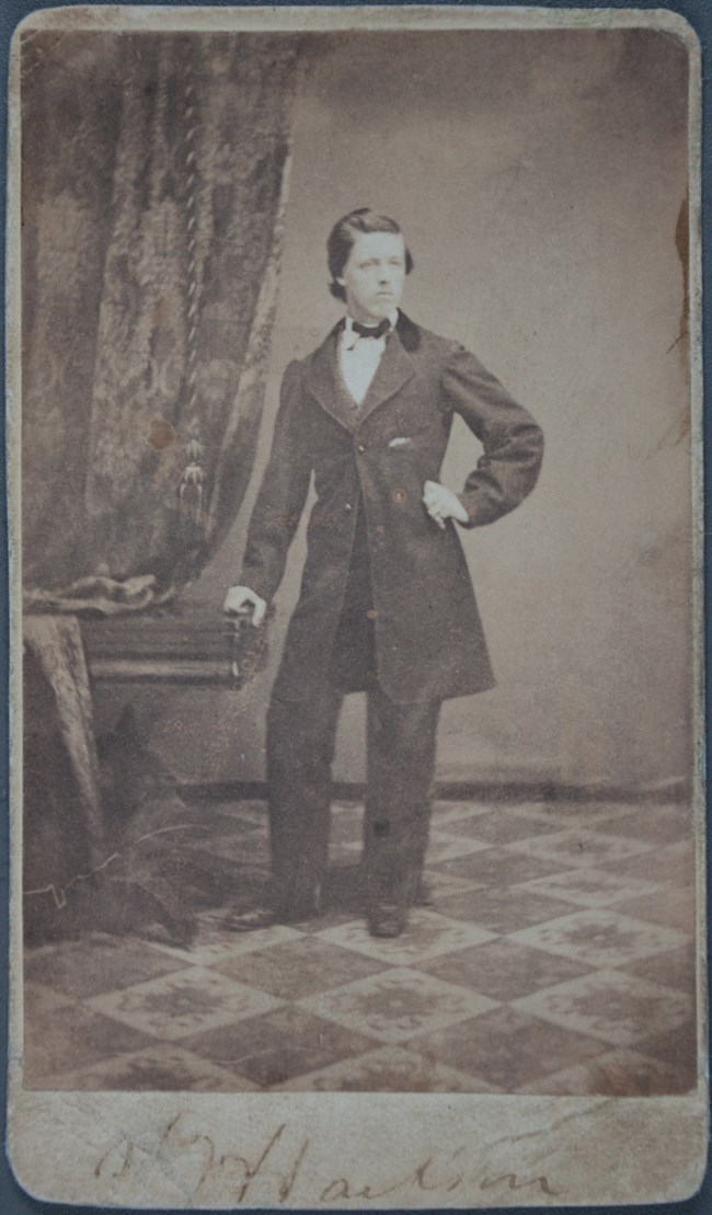 A young man wearing a suit stands with his right hand resting on a table.