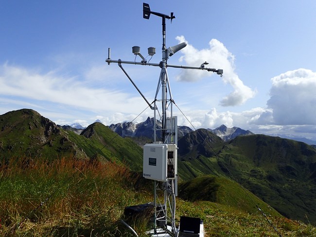 Deception Hills Weather Station Weather station sits on a green hilltop with rugged mountains behind.
