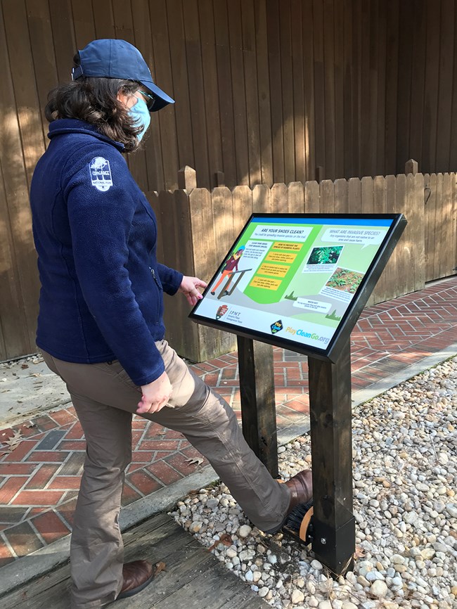 person stands outside reading a wayside exhibit and cleaning their shoes with the boot brush under the sign