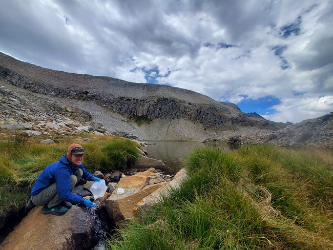 Woman crouches down on rocks near the outlet of a mountain lake and collects water samples in a plastic jug..