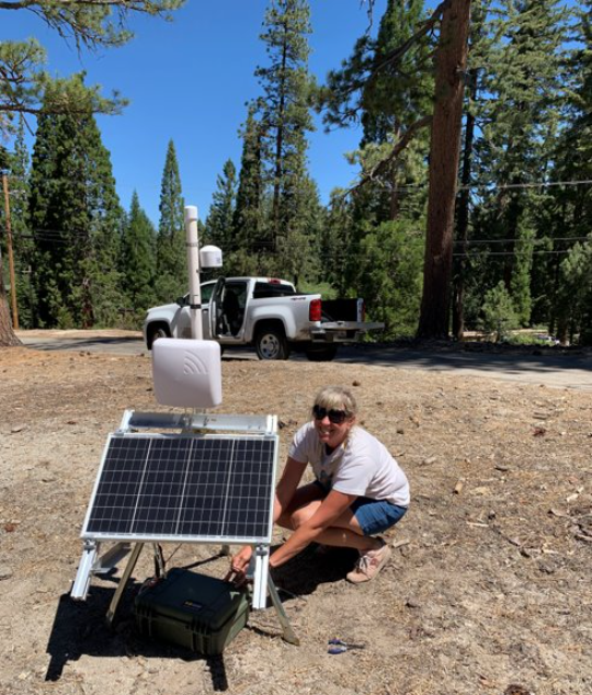 SEKI Purple Air Ariane (Ari) Sarzotti, SEKI Smoke and Weather Technician, deploying a portable PA system with solar panel on level bare ground with white work truck and evergreen trees in background.