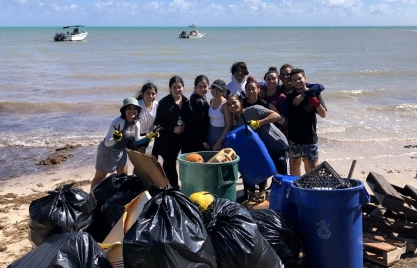 A group of volunteers poses behind a pile of collected debris and trash bags on the beach, the ocean stretches behind them, two boats sit on the water.