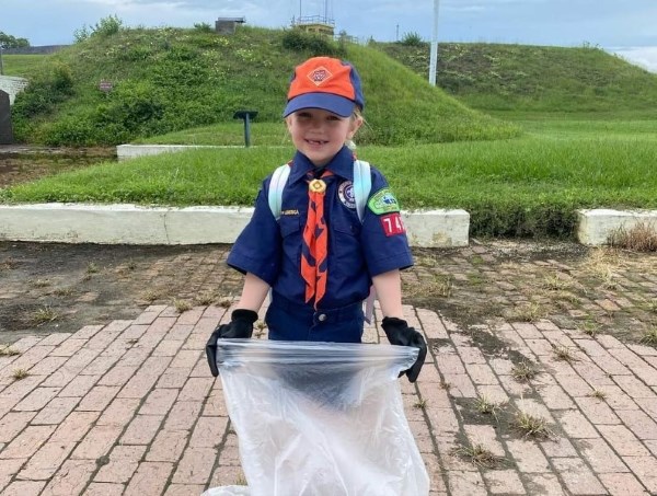A young girl poses for a picture in her Cub scout uniform, she wears gloves as she holds out a trash bag.