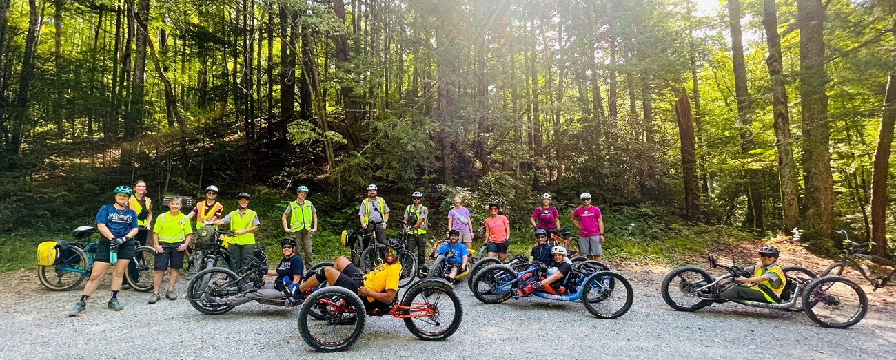 A group of people on adaptive bikes and others standing in front of a trees in the sun.
