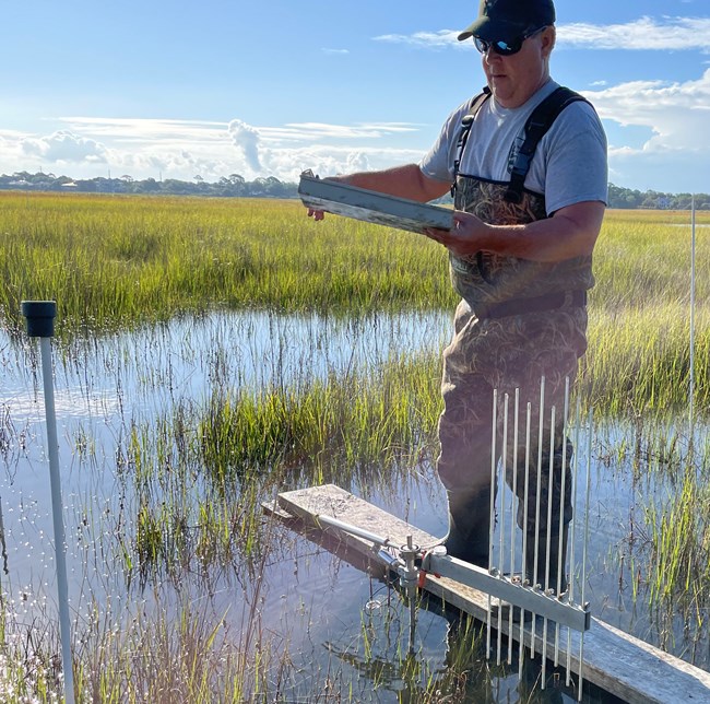 SET Data Man holding a metal box, standing on a platform in a marsh with metal pipes coming out of the marsh
