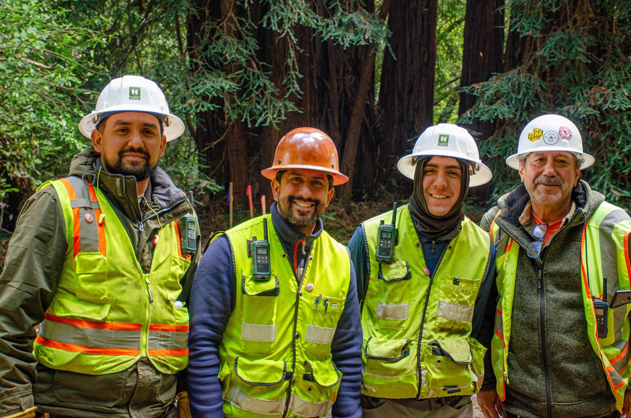 Four men wearing hard hats and flourescent yellow safety vests stand together and smile for a photo among the redwoods of Muir Woods.