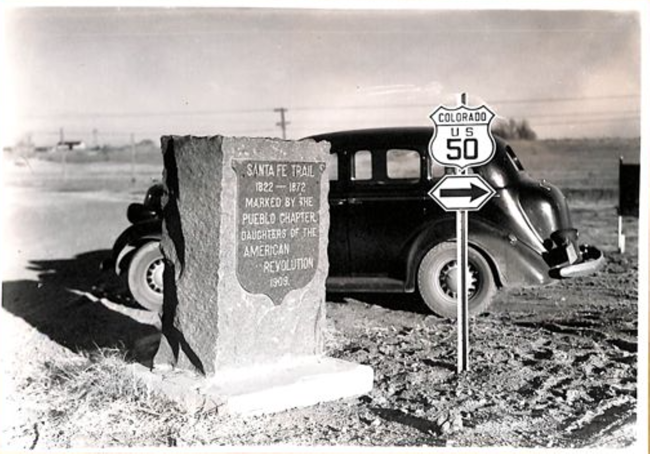 Stopping to understand the crossroads of history Black 1930s vehicle parks by a Santa Fe Trail marker and sign for Colorado Hwy 50