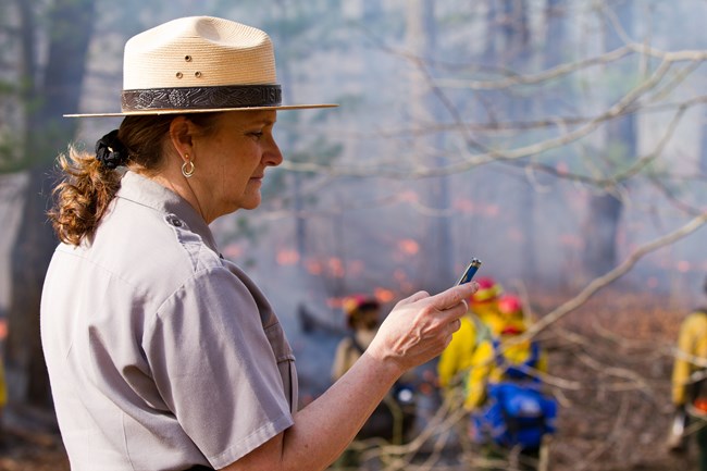 A park ranger checks her cell phone while wildland firefighters work in the background.