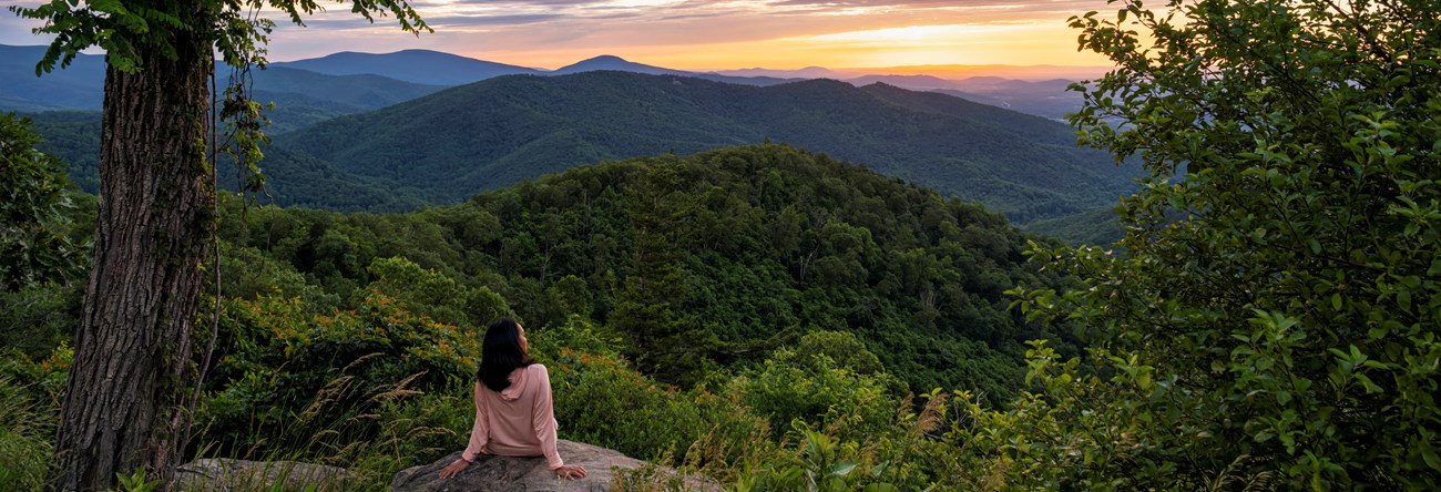 A visitor sitting on an overlook with a pink and purple sunset over wooded mountains.