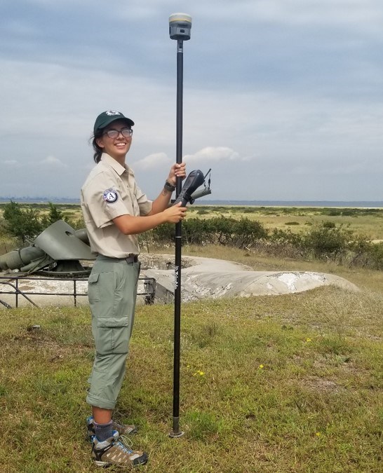 intern stands near the shore holding a tall GPS receiver