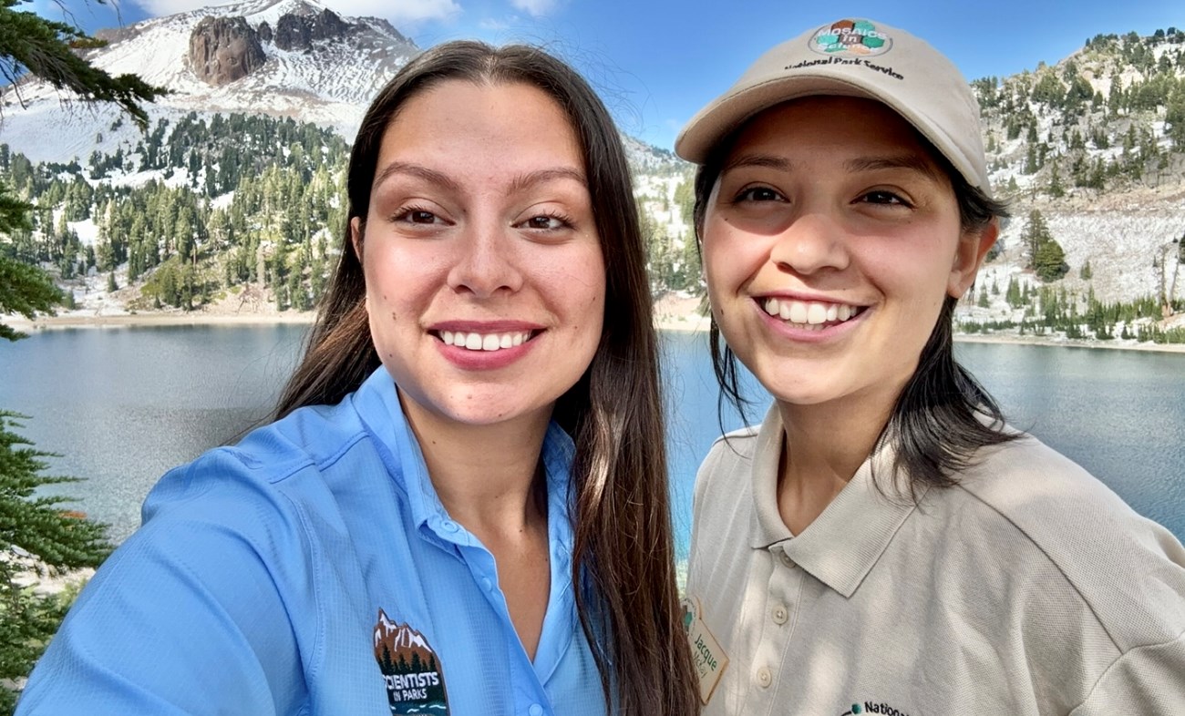 SIPMelissaDeLeon+MISJacqueMcKay+AtLAVO(selfie) Selfie of two women smiling side by side in front of a mountain lake surrounded by snow-covered slopes.