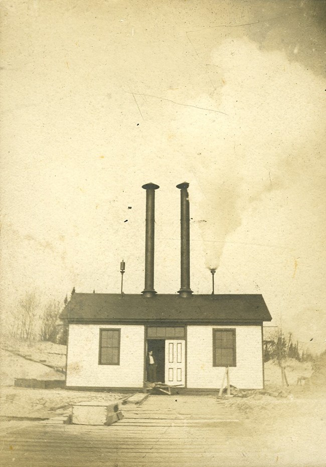 South Manitou Island's fog signal building. An aged black and white photo of a small rectangular building with a person leaning in the door frame. Two small cylindrical tubes and two large cylindrical tubes stick out of the roof.