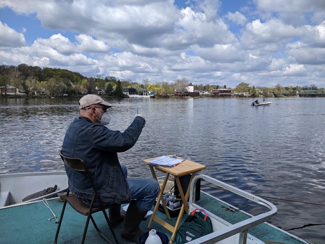 A citizen scientist in mask sits on the edge of the floating classroom on a wide river taking measurements