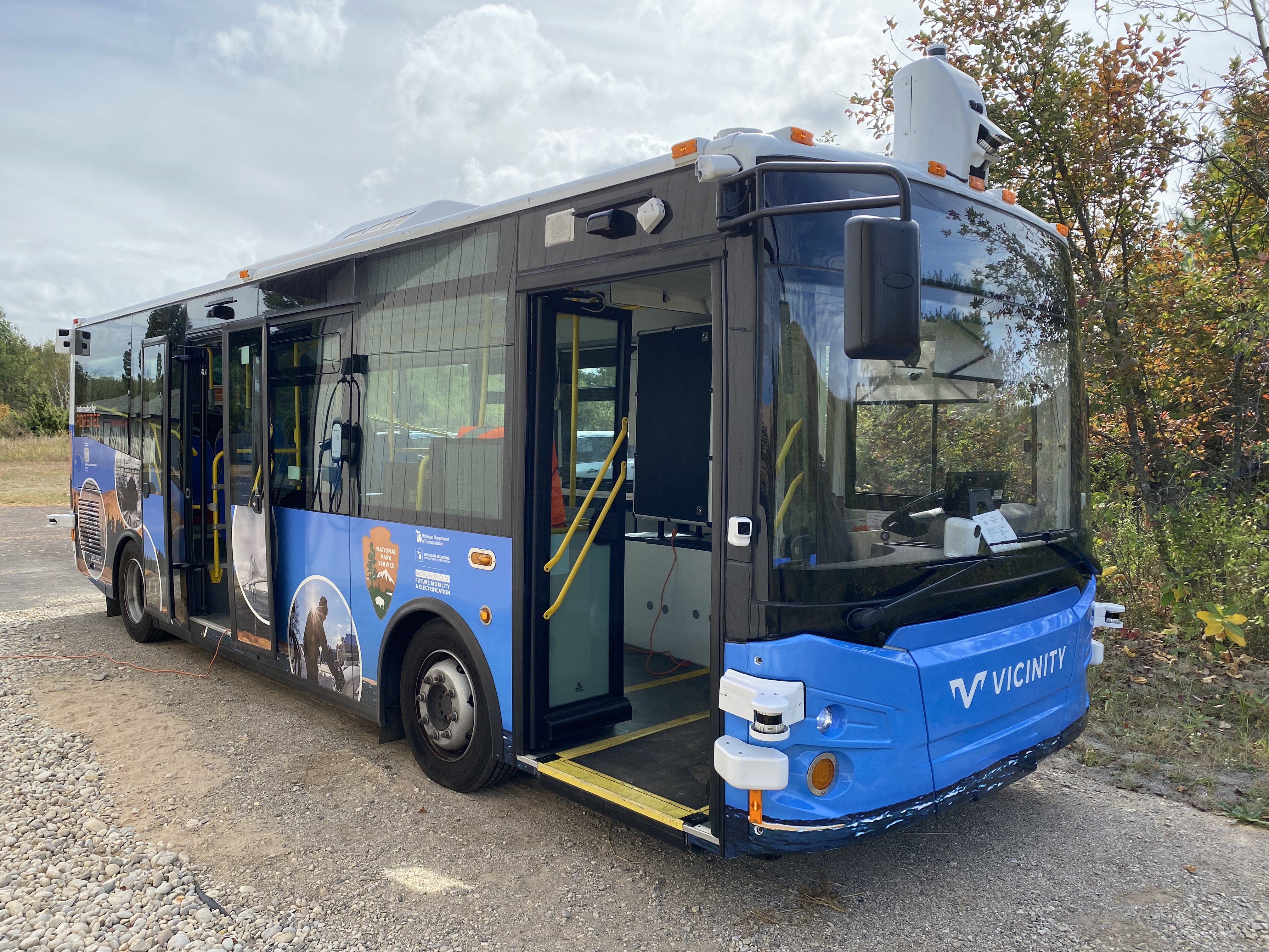 Blue and black electric bus is parked in front of small trees in a gravel parking lot