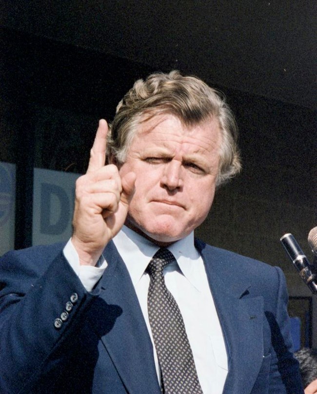 A color photo of Ted Kennedy behind a podium with his index finger raised