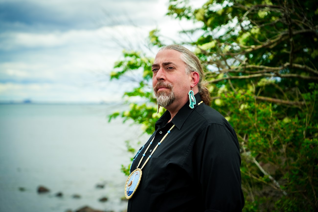 SZimmerman Headshot Headshot of a person wearing beaded earrings and a medallion with crane motif in front of a lake.