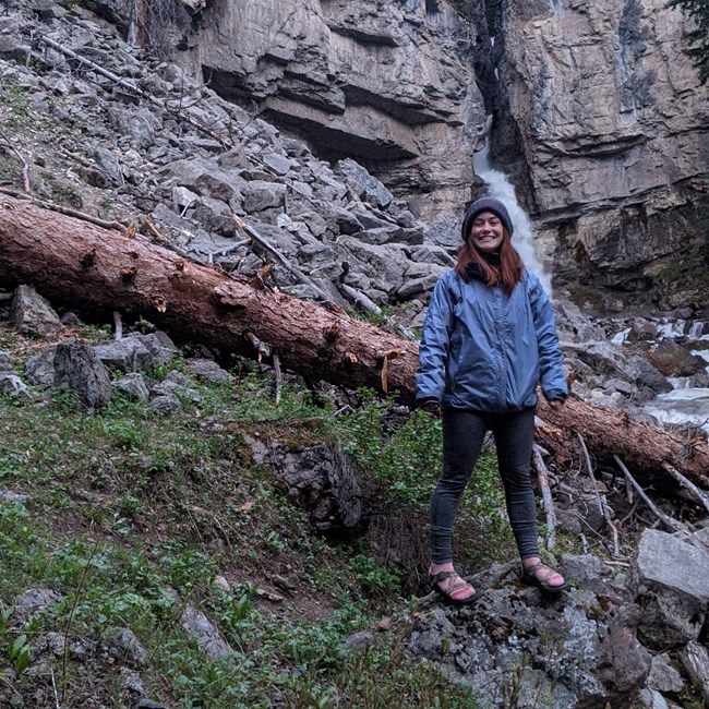S Milbauer Woman stands on rocky slope with a waterfall streaming from a cliff behind her.
