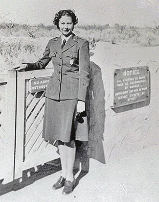 A woman wearing an NPS uniform with her badge pinned to her left side chest pocket is seen with her right arm on a gate. She faces left and holds sunglasses in her left hand.