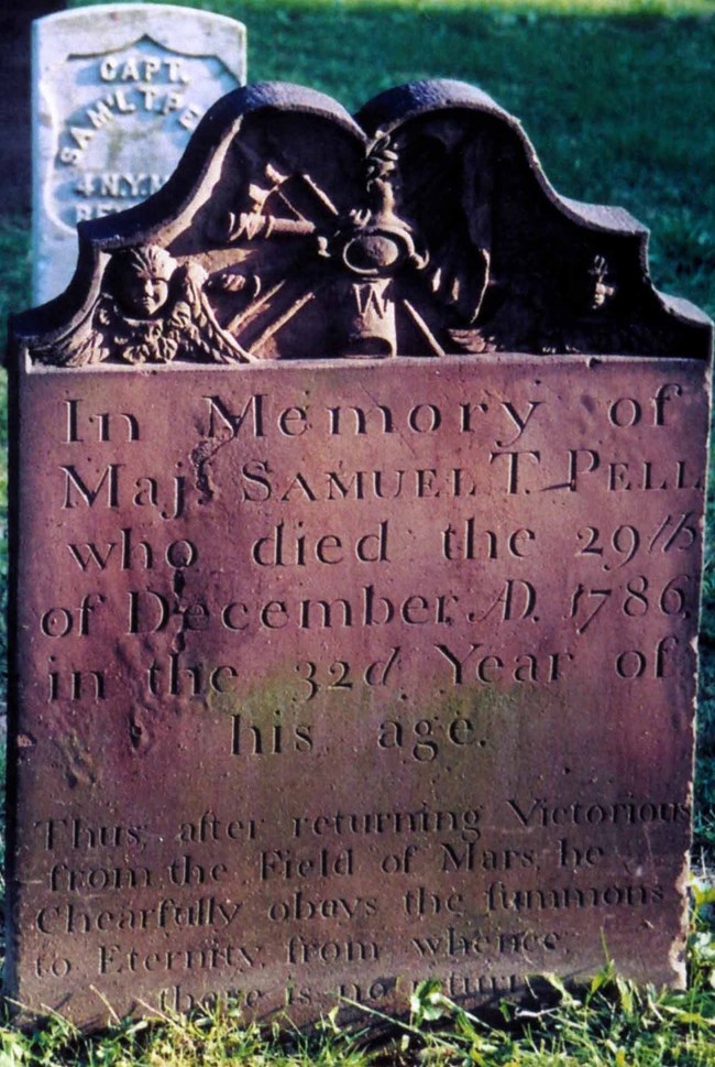 Reddish brown grave marker, with inscription, and designed carving at the top, set in a cemetery