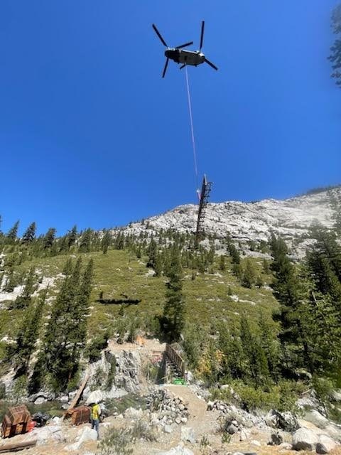 San Joaquin Bridge component being flown in. A helicopter hovers overhead with a piece of construction material for the San Joaquin Bridge hangs from a tow rope below it.