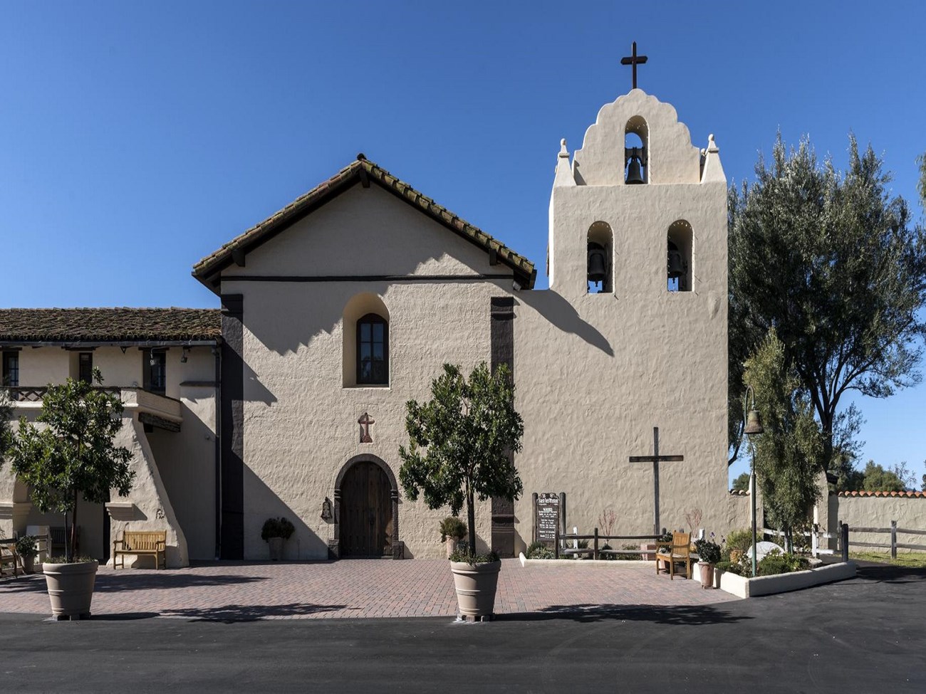 Santa Ines Mission Exterior Tan building, shorter on the left, taller in the middle, and a bell tower to the right. Roof and doors are brown. There are trees in the foreground.