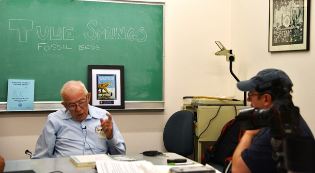 Santucci_figure_2_Vance_Haynes Photograph of 2 people seated at a table. Chalboard behind the interviewee says "Tule Springs Fossil Beds"