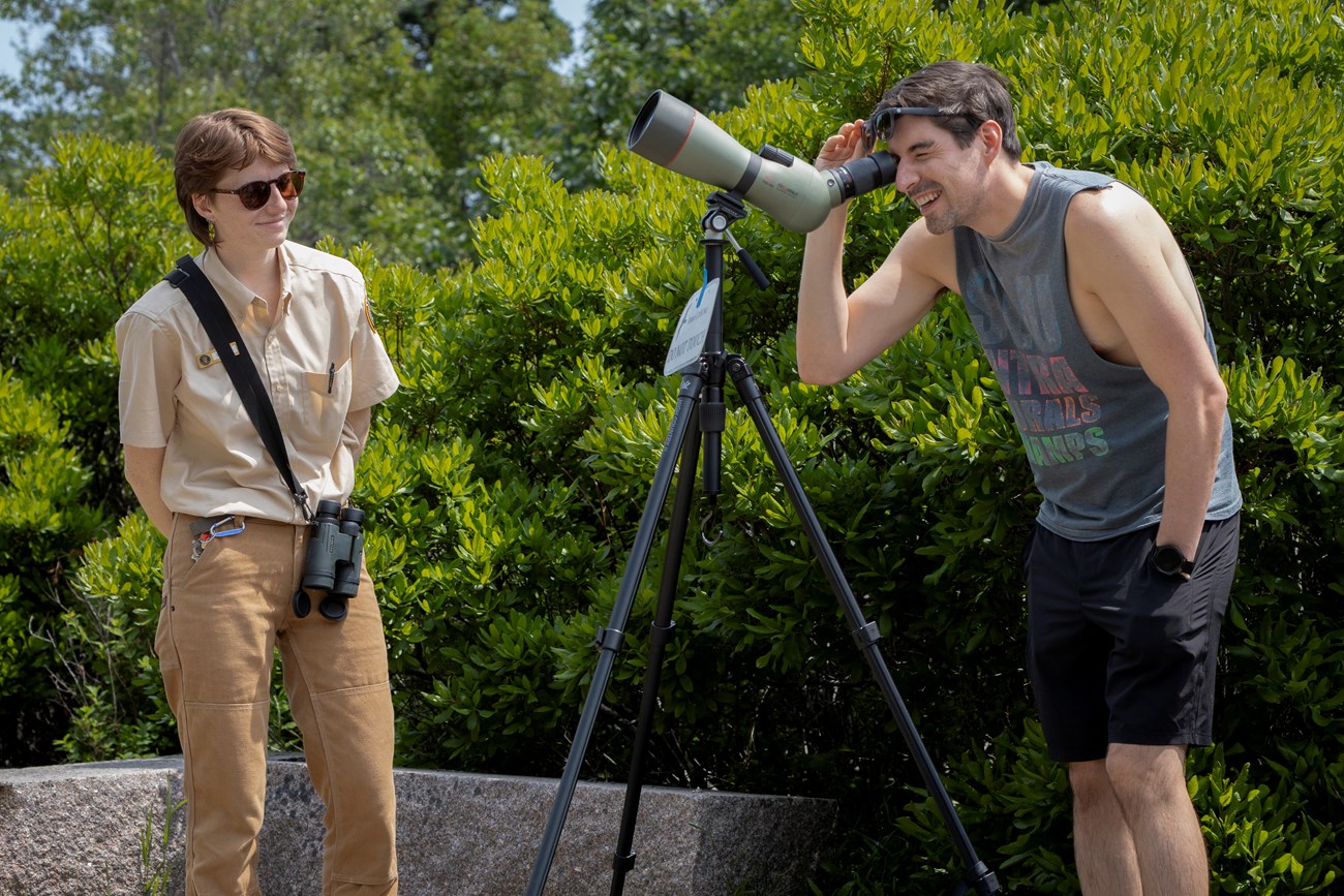 Sara talking to a visitor who is looking through a spotting scope pointed at a nesting peregrine falcon chick on the Precipice Trail. Intern talking to a visitor who is looking through a spotting scope pointed at a nesting peregrine falcon chick on the Precipice Trail.