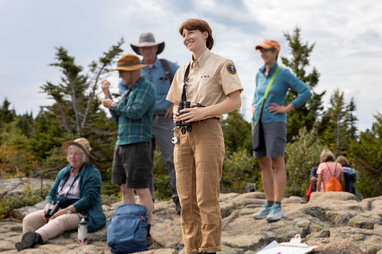 Sara York, Raptor Intern, watches migrating raptors on Cadillac Mountain with visitors. Raptor intern holds a pair of binoculars while smiling as she watches migrating raptors on Cadillac Mountain with visitors.