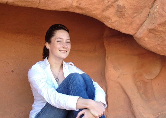Woman sitting under desert redrock overhang smiling with her arms wrapped around her knees.
