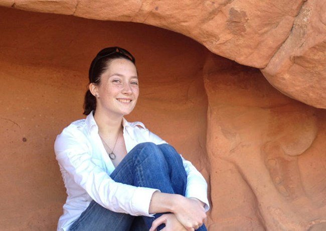 Sarah Wright Woman sitting under desert redrock overhang smiling with her arms wrapped around her knees.